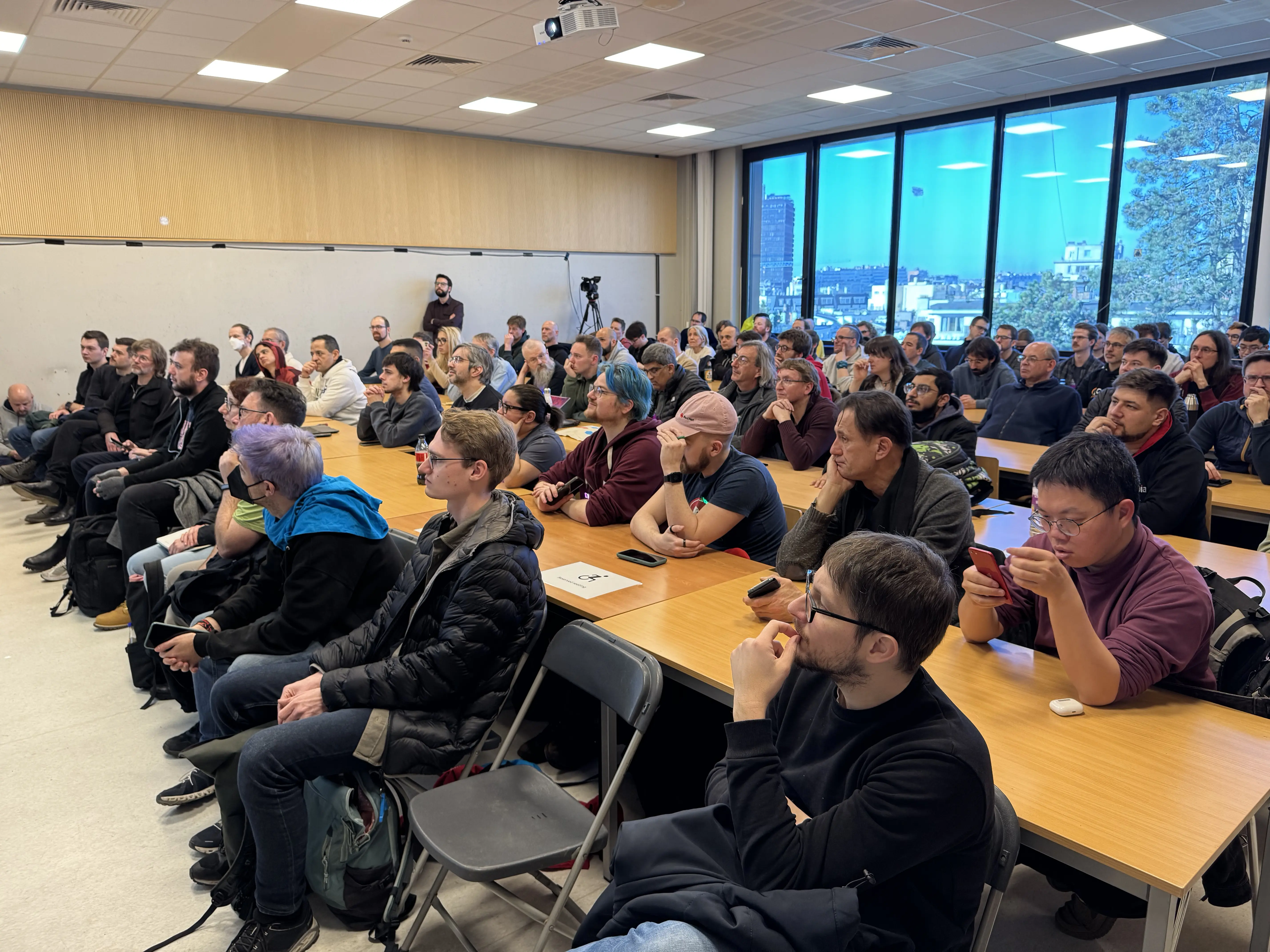 A picture of people sitting in a university lecture room. They are listening to a speaker off the frame. The room is almost full.